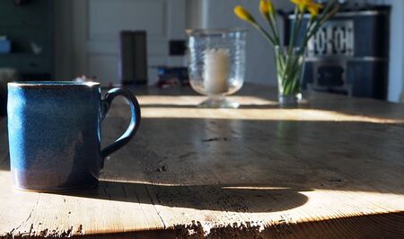 Blaue Tasse auf altem Holztisch, Sonnenstrahl von links, Kerze in einem Glasvase und  eine Vase mit Osterglocken. Kachelofen im Hintergrund rechts.