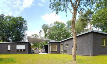 Flaches graues Kitagebäude in einer grünen, von Bäumen umgebenen Wiese unter blauem Himmel.